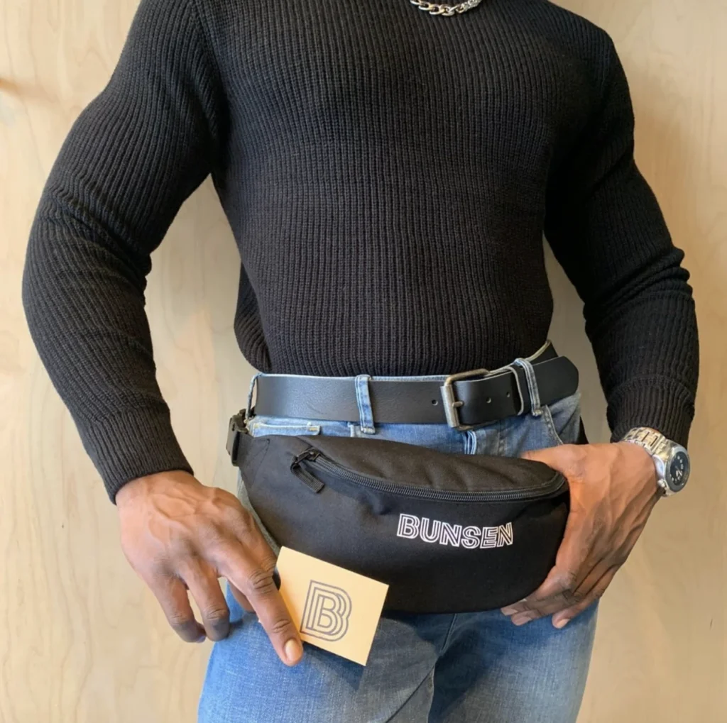 Person wearing a black jumper and jeans, holding a Bunsen branded belt bag.